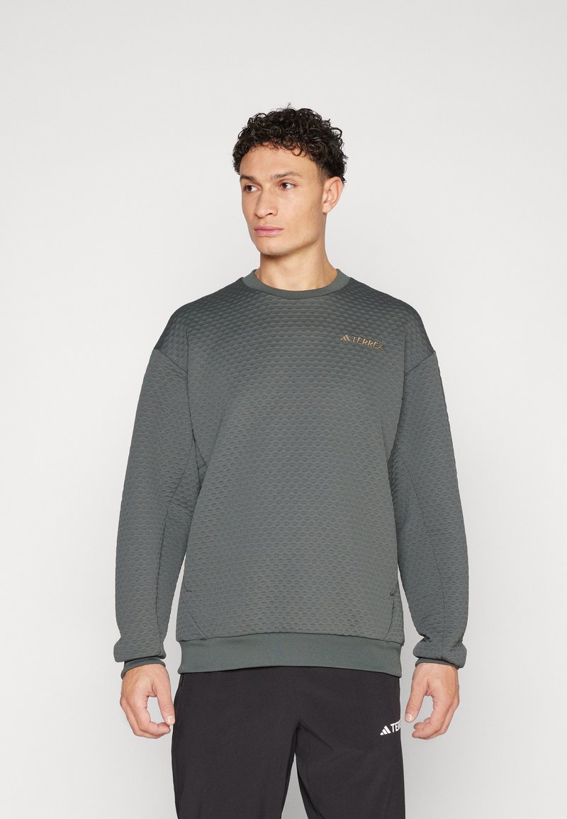 Sweatshirt texturé vert foncé avec un col rond et des poignets côtelés, présentant un petit logo doré sur le haut de la poitrine gauche.