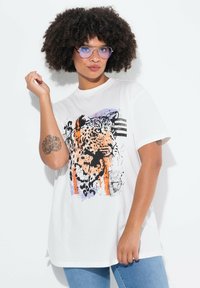 Bílý oversized tričko s leopardím potiskem v oranžové, černé a fialové barvě, s krátkými rukávy a kulatým výstřihem.