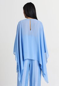 Poncho azzurro chiaro, semi-trasparente, con una vestibilità morbida, caratterizzato da un collo rotondo e un dettaglio a chiave sulla schiena. Tessuto morbido con maniche fluide.