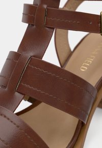 Καφέ δερμάτινο σανδάλι με σταυρωτούς ιμάντες, λεία επιφάνεια και χαμηλό τακούνι. Το εσωτερικό είναι ανοιχτό μπεζ με ανάγλυφη επωνυμία.