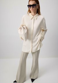 Chemise en soie couleur crème avec un col texturé, associée à un pantalon flared beige clair et des escarpins noirs à bout pointu. Le mannequin tient une pochette.