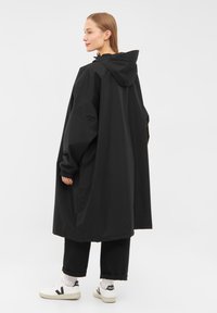 Black waterproof coat with a hood, oversized design, elastic cuffs, and side pockets, paired with black pants and white sneakers.