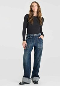 Jeans de mezclilla azul deslavado con un ajuste holgado, que cuentan con grandes bolsillos frontales y dobladillos remangados. Combinados con una blusa negra de manga larga.