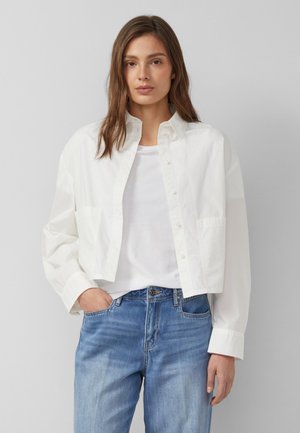 Jeune femme portant une chemise blanche ample et courte à boutons par-dessus un t-shirt blanc et un jean bleu clair, debout devant un fond gris.