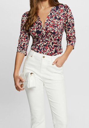 Kvinde iført en rød, navyblå og beige mønstret ruchet top med trekvartlange ærmer, hvide bukser, og holder en hvid quiltet clutch-taske.