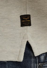 Beige textured top with a label featuring "PME LEGEND," showing cream seams and a split hem, layered over dark denim pants.