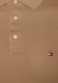 Polo shirt beige en tissu texturé, doté d'une patte de boutonnage à trois boutons et d'un petit logo brodé en rouge, blanc et bleu.