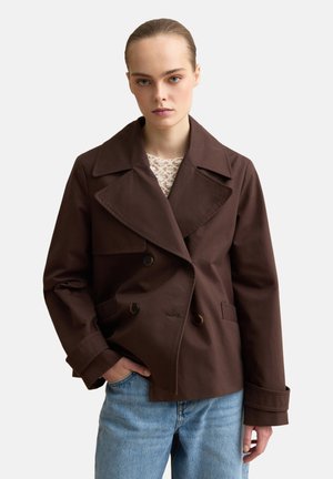 Jonge vrouw met naar achteren gestylede haren, gekleed in een donkerbruine dubbelgeknoopte jas en lichtblauwe spijkerbroek, staand met één hand in de zak.