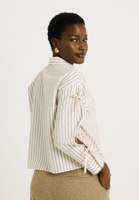 Femme portant un chemisier rayé beige et blanc avec des nœuds sur les côtés des manches, associé à un pantalon beige, se retournant par-dessus son épaule.