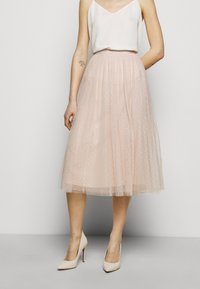 Jupe en tulle rose pâle à motif à pois, longueur mi-mollet, design plissé, associée à un haut crème et des talons hauts couleur nude.