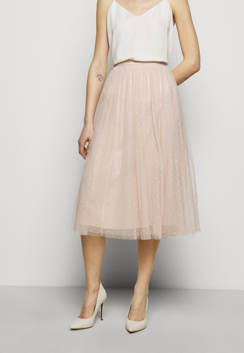 Jupe en tulle rose pâle à motif à pois, longueur mi-mollet, design plissé, associée à un haut crème et des talons hauts couleur nude.