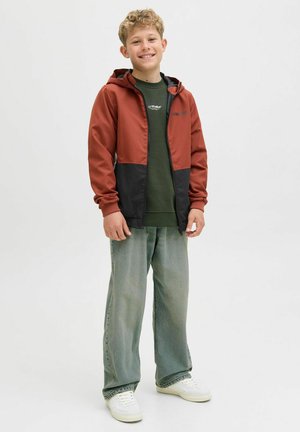 Veste zippée rouge et noire avec une capuche, en tissu lisse ; associée à un sweat-shirt vert olive et un jean bleu clair. Baskets blanches.