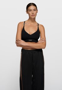 Top corto negro con diseño cruzado, con tirantes finos y una banda con logo. Combinado con pantalones negros, delineados con rayas marrones.