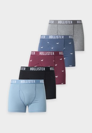 Noormees seisab paljajalu, kandes halli Hollisteri bokserit valgel taustal.