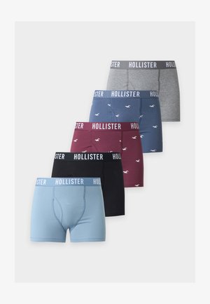 Noormees seisab paljajalu, kandes halli Hollisteri bokserit valgel taustal.