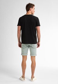 T-shirt in cotone nero, pantaloni corti con orlo arrotolato verde menta e sneakers di colore chiaro, visti da dietro su uno sfondo semplice.