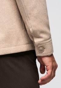 Giacca in lana beige con superficie texturizzata, dotata di polsini con bottone e orlo dritto, indossata sopra pantaloni scuri.