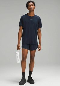 Maglietta e pantaloni sportivi blu navy in tessuto leggero, accompagnati da una bottiglia d'acqua bianca con un logo. Sneakers nere completano l'outfit.