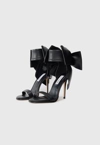 Black high-heeled sandals with a wide ankle strap featuring a decorative bow. Made of smooth leather, with a sleek minimalistic design.