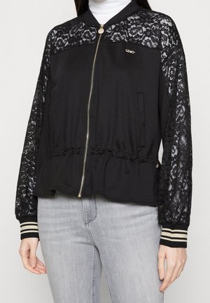 Femme portant une veste noire zippée avec des manches et des épaules en dentelle, des poignets côtelés avec des rayures blanches, assortie à un jean gris clair.