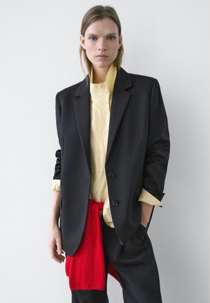 Frau trägt einen schwarzen Blazer über einem blassgelben Hemd mit Knöpfen und einem roten Pullover, der um die Taille gebunden ist, und steht vor einem weißen Hintergrund.