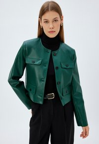 Giacca in pelle verde con collo rotondo, chiusura frontale con bottoni, due tasche sul petto e design strutturato, indossata sopra un dolcevita nero.