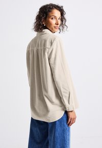Vrouw met krullend haar, gekleed in een beige corduroy shirt en blauwe spijkerbroek, kijkt over haar schouder tegen een effen witte achtergrond.