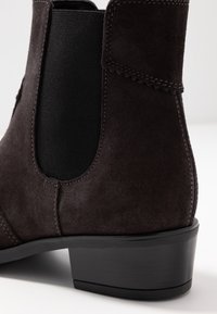 Donkerbruine suède enkellaars met een gestructureerde afwerking, elastische zijpanelen en een lage zwarte hak voor stabiliteit. Gestikte accenten zichtbaar.