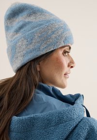 Berretto lavorato a maglia in blu chiaro e grigio con una texture morbida. La persona indossa una giacca blu con un alto colletto e dettagli visibili della cerniera.