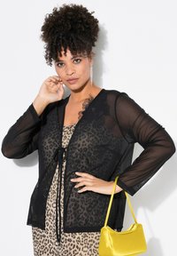 Cardigan in rete nero con nodo frontale, caratterizzato da dettagli in pizzo. Indossato sopra un vestito in stampa leopardata, abbinato a una borsa in satin gialla.