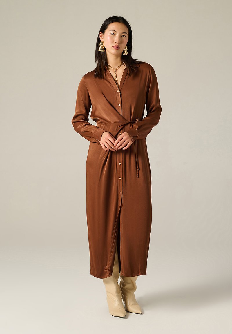 Femme portant une longue robe brune boutonnée avec une ceinture à la taille, des bottes beiges et de grandes boucles d'oreilles en or, se tenant devant un fond beige uni.