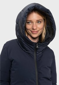 Donkerblauwe pufferjack met een grote, capuchon kraag, ritsluiting en een gladde, glanzende textuur. Beschikt over subtiele stikdetails en korte mouwen.