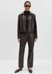 Braune Kunstleder-Cropped-Jacke mit zwei Vorder-Taschen, kombiniert mit passenden braunen Hosen und spitzen Mules. Die Accessoires umfassen schwarze Sonnenbrillen.