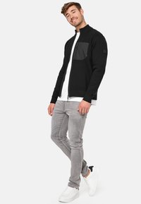 Sort zip-up sweater med en tekstureret sort brystlomme, slanke grå jeans og hvide sneakers. Modellen poserer foran en ensfarvet baggrund.