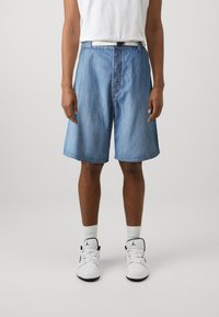 Egy férfi világoskék farmernadrágot, fehér zoknit, fekete-fehér magasszárú tornacipőt és fehér pólót visel, szürke háttér előtt.