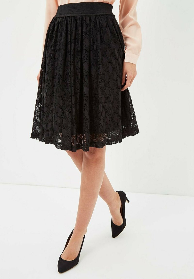 Falda de encaje negro con un patrón de diamantes texturizado, diseño plisado y cintura elástica, combinada con tacones altos negros.