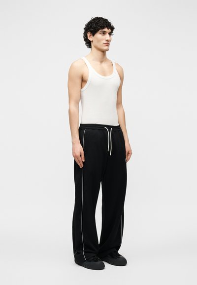 Νέος άνδρας με σγουρά μαλλιά, φοράει λευκή φανέλα και άνετο μαύρο παντελόνι με λευκή λεπτομέρεια, στέκεται μπροστά σε απλό ανοιχτό φόντο.