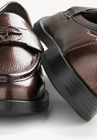 Brune læderloafer med en struktureret overflade, der har en sølvaccent på stroppen og en sort gummisål. Nærbillede.