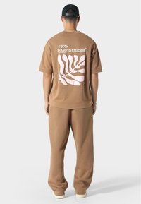 Ruskea ylipitkä t-paita ja yhteensopivat collegehousut, joissa on suuri vaaleanpunainen lehtigrafiikka ja "IRASUTO STUDIOS" -teksti selkäpuolella. Pehmeä kangas.