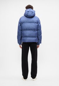 Giacca a vento blu con cappuccio, caratterizzata da sezioni trapuntate, una texture liscia e polsini elasticizzati, abbinata a pantaloni neri e sneakers chiare.