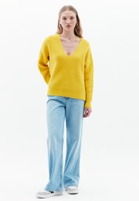 Geel gebreide trui met een diepe V-hals, gestructureerde diagonale strepen en verlaagde schouders, gecombineerd met lichtblauwe flared jeans en grijze sneakers.