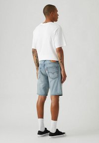 Weißes T-Shirt mit kurzen Ärmeln, hellblaue Jeans-Shorts, schwarze Sneakers, weiße Socken. Rückansicht zeigt lässigen Streetwear-Stil und Tattoos.