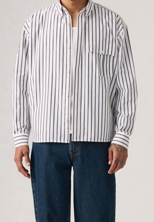 Homme portant une chemise blanche à boutons avec des rayures verticales noires sur un sous-vêtement blanc, associée à un jean bleu foncé.