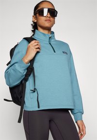 Himmelblaues Pullover-Sweatshirt mit hohem Kragen und halbseitigem Reißverschluss. Schwarzes Logo auf der Brust. Sichtbare schwarze Rucksackträger. Dunkle Leggings getragen.