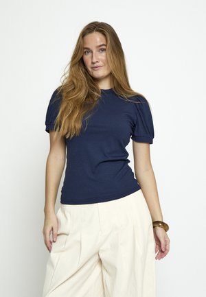 Žena s dugom svijetlosmeđom kosom, odjevena u tamnoplavu majicu kratkih rukava i široke, visokog struka kremaste hlače, na jednobojnoj bijeloj pozadini.