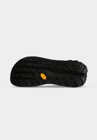 Semelle de chaussure en caoutchouc noir avec des rainures profondes et un petit logo Vibram jaune au centre sur fond blanc uni.