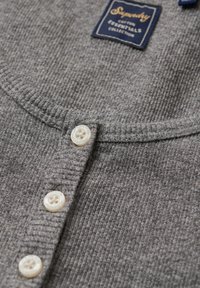 Grå ribbet bomuldsstriketop med rund halsudskæring, prydet med tre hvide knapper og et blåt mærke, der står "Superdry Essentials Collection."