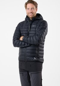 Homme portant une doudoune noire à capuche Swedemount, bras croisés, pantalon sombre de plein air, souriant, sur fond blanc.