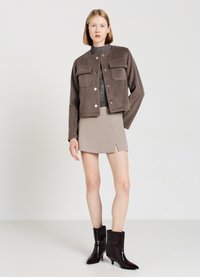 Veste en velours côtelé marron avec des boutons dorés et deux poches poitrine, associée à une mini jupe beige comportant une fente, et des bottines noires brillantes.