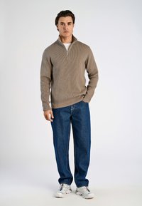 Beige gerippter Reißverschluss-Pullover, blaue locker sitzende Jeans und weiße Sneaker mit bunten Akzenten, die vor einem einfarbigen Hintergrund stehen.